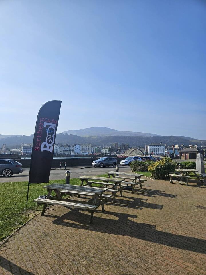 The Northern Pool Cafe flag beside the picnic benches on Mooragh Promenade, with Ramsey harbour and the hills behind
